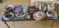 Full view of bakeware laid out on table showing assortment of glass dishes in clear and purple tint with some covered casseroles and mixing bowls.