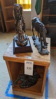 Bronze-tone End of the Trail replica statue and metal horse statue on small wooden end table. Metal ship statue on shelf below.