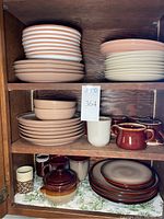 Photo of various stacked plates in different colors and bowls and mugs placed on wooden shelves. Shows ivory and spruce colored pieces.