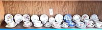 Wide shot of 18 teacups and saucers arranged on a wooden shelf, showing various floral patterns and colors including blues, purples, yellows, and pinks.
