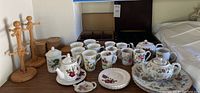 Overall view of bone china teacups, small teapots with floral designs, cookie plates, wooden tea racks holding cork coasters, and other small accessories.