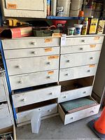 White drawer unit with multiple labeled drawers, some open showing various supplies inside such as sandpaper and gardening hardware. Drill box and tools visible on top.