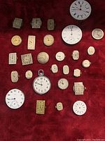 Full view of 26 antique watch dials and movements placed on a red cloth background showing various shapes and sizes.