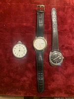 Three vintage watches laid on red fabric including a silver-tone pocket watch, gold-tone wristwatch with black leather strap, and a black dial wristwatch with brown leather strap.