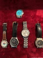 Top view of five wrist watches arranged on red background. Various styles including leather and metal bands, different dial colors and designs.
