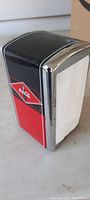 Side angle showing the black top and red front with Coca-Cola diamond logo and bottle graphic with chrome trim, holding paper napkins.