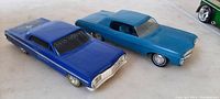 Two hard plastic car models, blue 1960s promotional car with MAD HOPPER decal and teal 1964 Chevrolet model, displayed on tan surface showing front and side details.