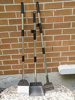 Three pooper scooper tools standing upright leaning against a brick wall. From left to right: flat spade, metal tray, and rake scooper with metal handles and black grips.