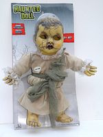 Front view of the haunted doll in original packaging, showing full doll in beige dress with greenish-gray belt and cracked face design.