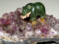 Close-up side view of the dark green polished jade bear holding a beige fish on a purple amethyst crystal cluster showing detail of the bear's body and fish mouth.