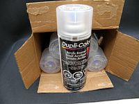 Six new unopened Dupli-Color Acrylic Enamel spray paint cans in original cardboard box, front view showing one can out of the box