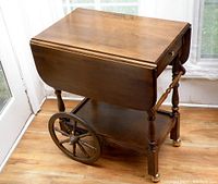 Vintage wooden tea cart with two drop leaf sides, medium brown finish, viewed from a corner angle showing large wooden spoked wheels and caster wheels