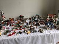 Overall view of the table with assorted Christmas village pieces, buildings, figurines, benches, bridges, trees, and lighthouse.