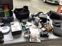 Table display showing Nikon digital camera with attached lens and extra zoom lens, Panasonic digital camera with flash, Panasonic portable CD player (blue and white), vintage Panasonic tape player, photo manuals, and various camera accessories