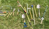 Full overview of the garden tools laid out on grass including shovels, rakes, blades, and other hand tools with wooden handles