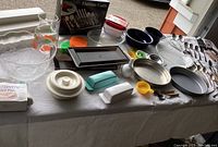 Photo showing a wide assortment of kitchen items including flatware, baking pans, glassware and storage containers laid out on a table.