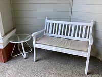 White 2-seater outdoor bench with beige cushion and white round side table with glass top placed on patio corner