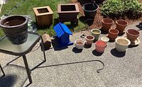Overview of lot showing various planters, plant stands, blue bird feeder, side table and garden accessories on concrete ground outdoors.
