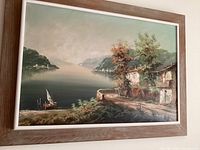 Framed painting showing cottage by the sea with boat and trees, wood frame with white trim.