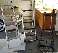 Photo showing two metal step ladders of different heights and one black metal hand truck.