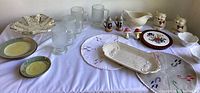 Wide view of three sets of vintage salt and pepper shakers, glass tumblers, hand painted plates, and serving pieces on white tablecloth