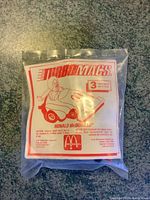 Front view of sealed package with red and white printed label showing Ronald McDonald driving a small toy car labeled Turbo Macs. McDonald's logo and caution information visible.