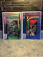 Photo shows both Green Lantern and Supergirl Futures End #1 comics side-by-side in protective cases. Green Lantern comic has a green and black holographic 3-D cover, and Supergirl comic has a blue and red holographic 3-D cover.