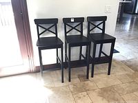 Three dark wood bar stools with X-back design positioned side by side near a door, showing their height and design details.