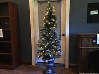 Full view of the artificial Christmas tree standing in decorative urn-style stand, lit with warm white lights.