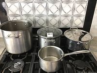 Four stainless steel pots and pans displayed on a stove: three stock pots of varying sizes and one small saucepan, two stock pots have lids (one glass, one metal).