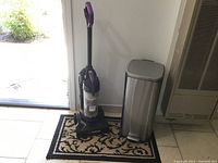 Photo showing upright Eureka Air Speed vacuum and stainless steel foot pedal trash can on a floor mat near an open door.