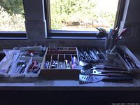 Wide photo of multiple trays and a wooden box containing various kitchen utensils and gadgets including knives, forks, spoons, peelers, tongs, spatulas and a mortar and pestle.