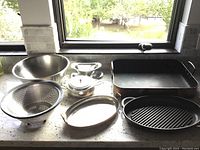 Image showing the various metal kitchen items including roasting pan, grill pan, gravy boats, mixing bowl, strainer, and covered serving dish on a countertop by a window.