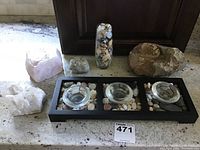 Overall view of collection including two crystals, a glass vase with stones, a large fossil rock, small stone and black candle holder tray with glass cups surrounded by stones.