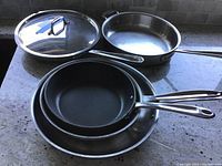 Photo showing four cooking pans including a sauté pan with glass lid, two frying pans and one shallow pan, arranged on a granite countertop.