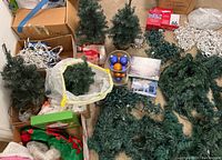 Four small artificial pre-lit Christmas trees, boxes containing string lights and ornaments, garland sections, and other Christmas decor laid out on the floor