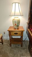 Lamp with Asian ceramic base and floral designs placed on wooden side table with turned legs and drawer.