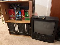 Sylvania TV/VCR combo unit next to two cardboard boxes containing VHS tapes of various brands and a remote control on top of the unit.
