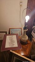 Photo of a handpainted floral table lamp on a wooden table with framed art pieces beside it.