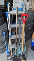 Lot overview showing the 2-way extension ladder, three shovels (including one with red handle), and shingle remover tools leaning against tires in a garage.