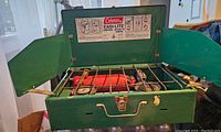 Green Coleman campstove open showing two red fuel burners, metal grates, and fold-out windshields with instructions inside lid.