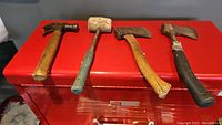 Four tools laid on a red surface: a claw hammer, mallet, two hatchets (one wooden handle, one black grip)