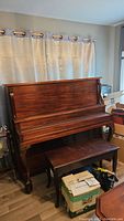 Full front view of upright piano with bench sitting underneath and boxes around the base.