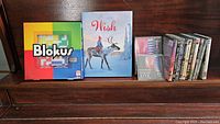 Image of Blokus board game, Wish book, 2 music CDs, and 6 DVDs neatly arranged on wooden surface.