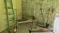 Wide view showing antique wooden bench with rusted metal frame, garden tools (pitchfork, shovel), and a green wooden ladder against stone wall on wooden floor.