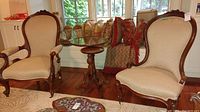 Two vintage beige upholstered parlor chairs with dark carved wooden frames, one with arms and one armless, displayed near a window with a wooden floor and a small round wooden table between them.