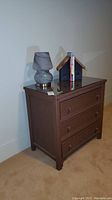 Front and angled view of 3-drawer brown wicker dresser with glass top, lamp, bookends, and books on top.