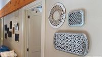 View of multiple vintage metal grates mounted on light colored walls, varying sizes and shapes with white and black finishes showing ornate openwork designs.