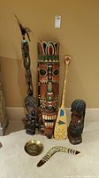 Photo showing all the items in the lot arranged on carpet: large colorful tribal mask, two wood carved statues, wooden paddle, painted boomerang, brass dish, and carved wooden staff.
