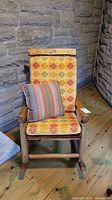 Front view of the natural wood rocker with cushions and striped throw pillow against stone wall